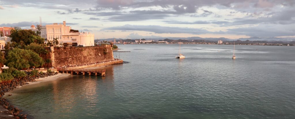 Old San Juan Bay taken from Castillo San Felipe del Morro, showcasing part of the shore during sunset with calm waters reflecting the golden light