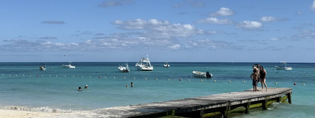 Pier extending into turquoise waters from the beach in Playa del Carmen, Mexico, with a family walking along the pier and boats visible in the background, showcasing a peaceful coastal scene