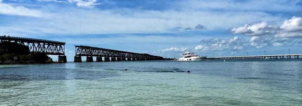 People snorkeling at Bahia Honda State Park under the bridge, with a large yacht visible in the background and the iconic bridge leading to Key West in the distance.