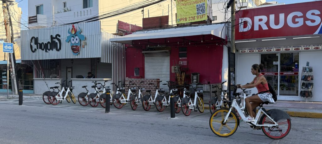 Bicycles lined up in front of stores in Playa del Carmen, with a woman riding by on one of the bikes, showcasing the ease of using the Biciplaya app for exploring the city