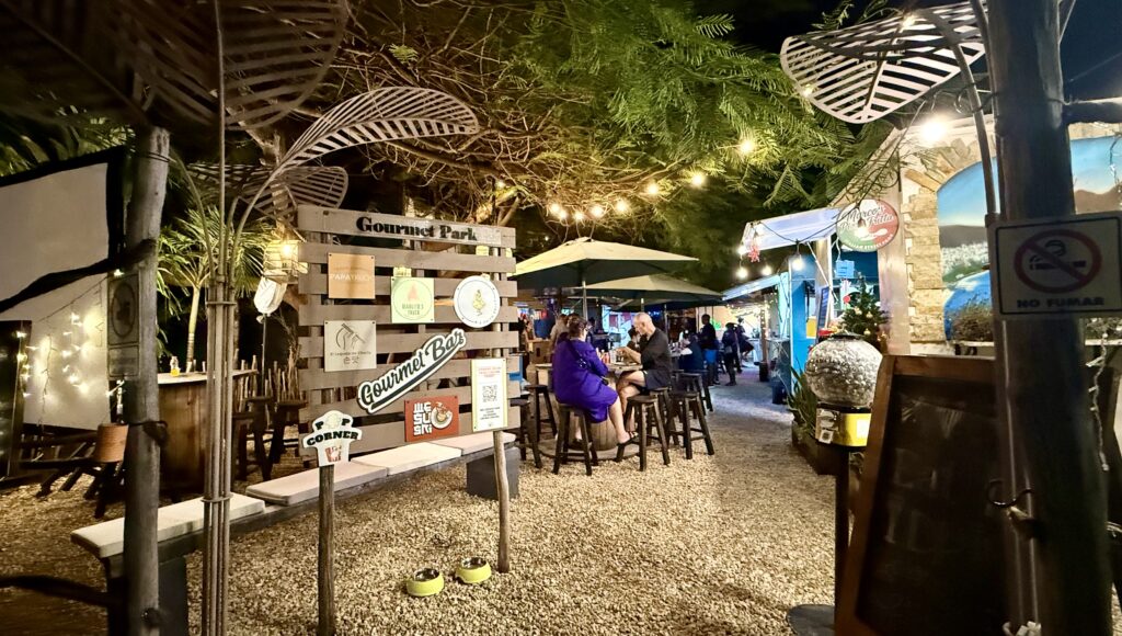 A colorful and vibrant sign for Gourmet Park in Playa del Carmen, Mexico, surrounded by tropical greenery and a warm, sunny atmosphere, inviting visitors to explore its culinary delight.