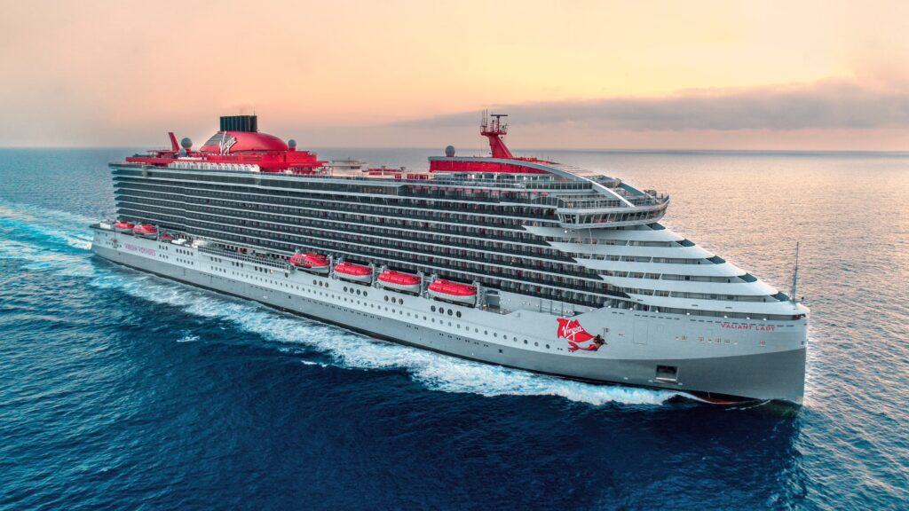 Virgin Voyages cruise ship sailing gracefully through the open ocean at sunset, with the sky painted in hues of orange, pink, and purple, creating a stunning backdrop of calm waves and the silhouette of the ship in the distance.