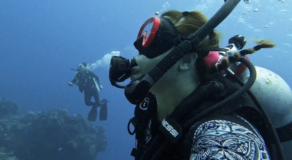 Scuba diving in Cozumel with crystal-clear waters and a fellow diver in the background, showcasing diving gear and the stunning underwater environment.