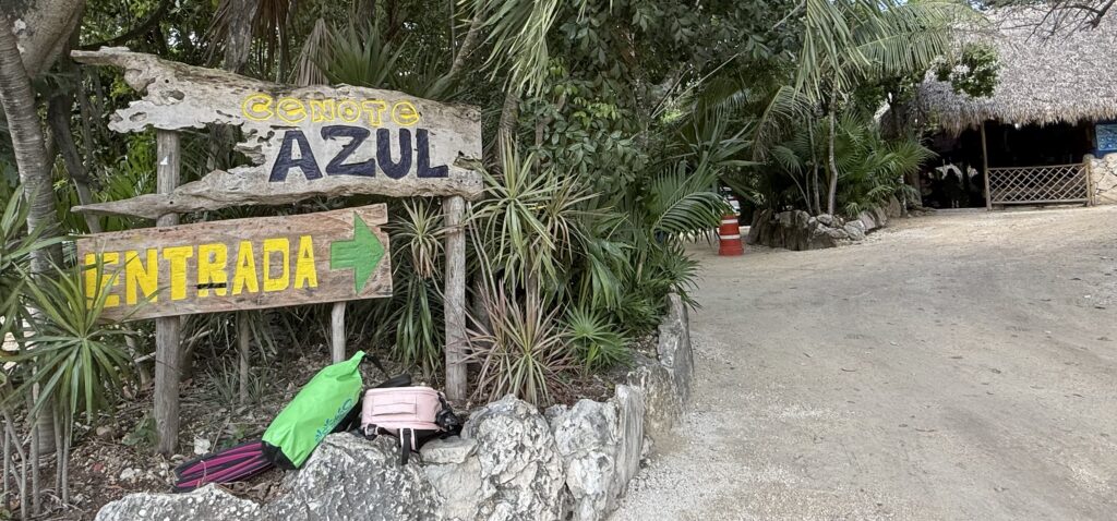 Entrance to Cenote Azul in Mexico, featuring gear and fins displayed on the wall alongside a sign reading 'Cenote Azulejo Entrada.' Surrounded by lush greenery, this serene spot offers a glimpse into the natural beauty of the cenote