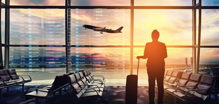 A traveler standing in the airport with luggage, watching a plane take off into the sunset with a vibrant sky in the background