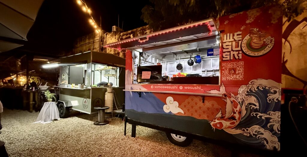 A vibrant sushi food truck with colorful signage, surrounded by greenery, serving fresh sushi dishes in Playa del Carmen. A lively outdoor dining space at Gourmet Park in Playa del Carmen with a DJ playing music in the background, creating a vibrant and fun atmosphere