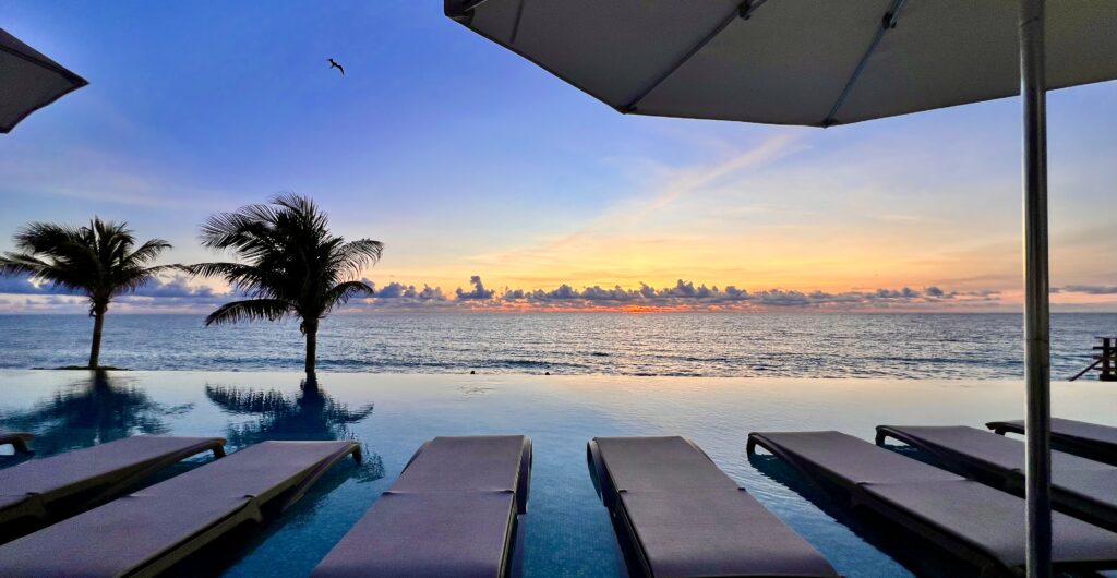 A picturesque view of the infinity pool at the all inclusive resorts Sun Palace Cancun, overlooking the tranquil Caribbean Sea as the sun rises, casting a serene glow across the water and creating a peaceful, luxurious atmosphere.