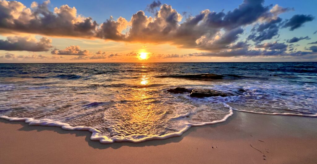 Waves crashing on the Caribbean shoreline at sunrise, with the tranquil reflection of the vibrant sky creating a stunning, peaceful scene.