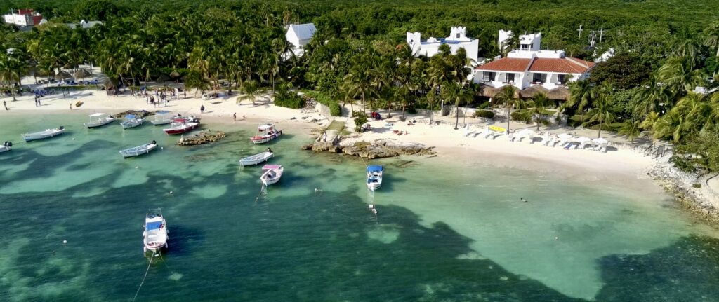 Drone view of Akumal beach, showcasing clear turquoise waters, surrounding boats, and beachfront buildings in the background for a picturesque coastal scene