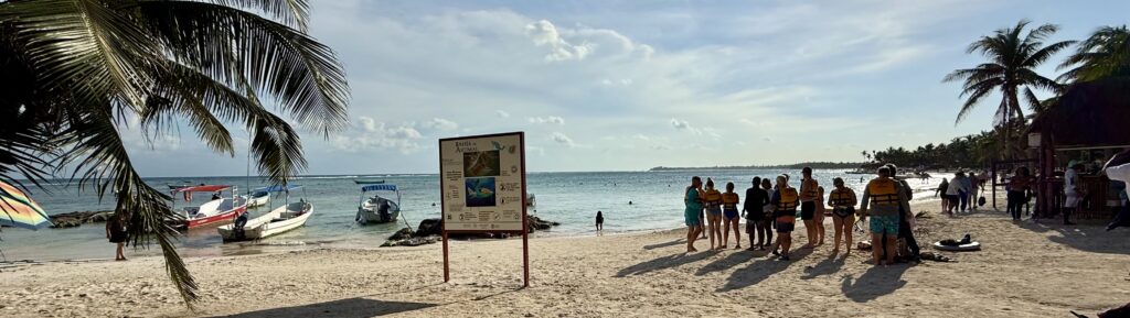 People lining up at the beach in Akumal for a swim with the turtles excursion, with boats and palm trees in the background creating a tropical setting