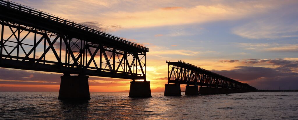 Bahia Honda Bridge at sunset, featuring vibrant hues of blue, yellow, and orange as the sun sets behind the bridge, creating a stunning, picturesque view with a break in the middle