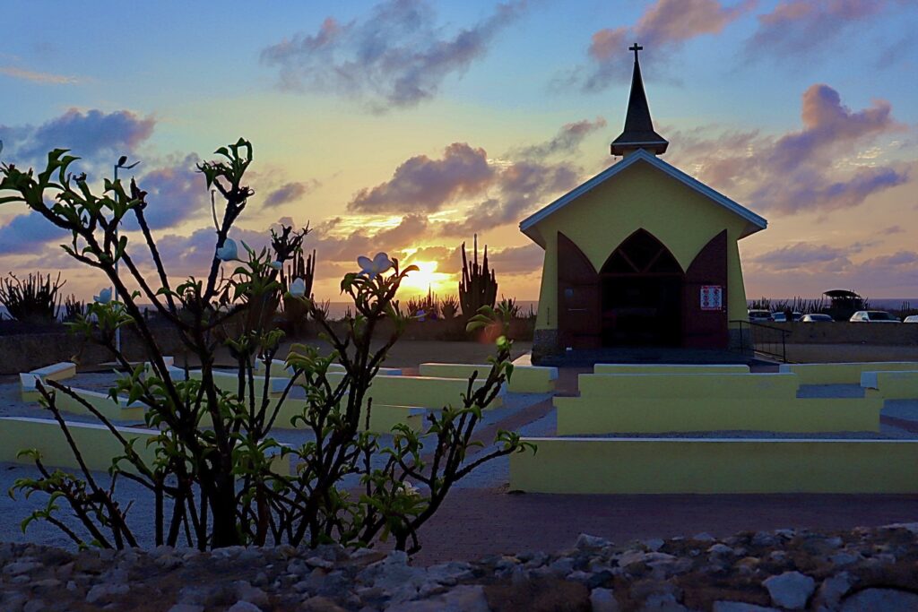 Beautiful sunrise at Alto Vista Chapel in Aruba, featuring vibrant flowers and towering cacti surrounding the historic church, creating a serene and picturesque setting